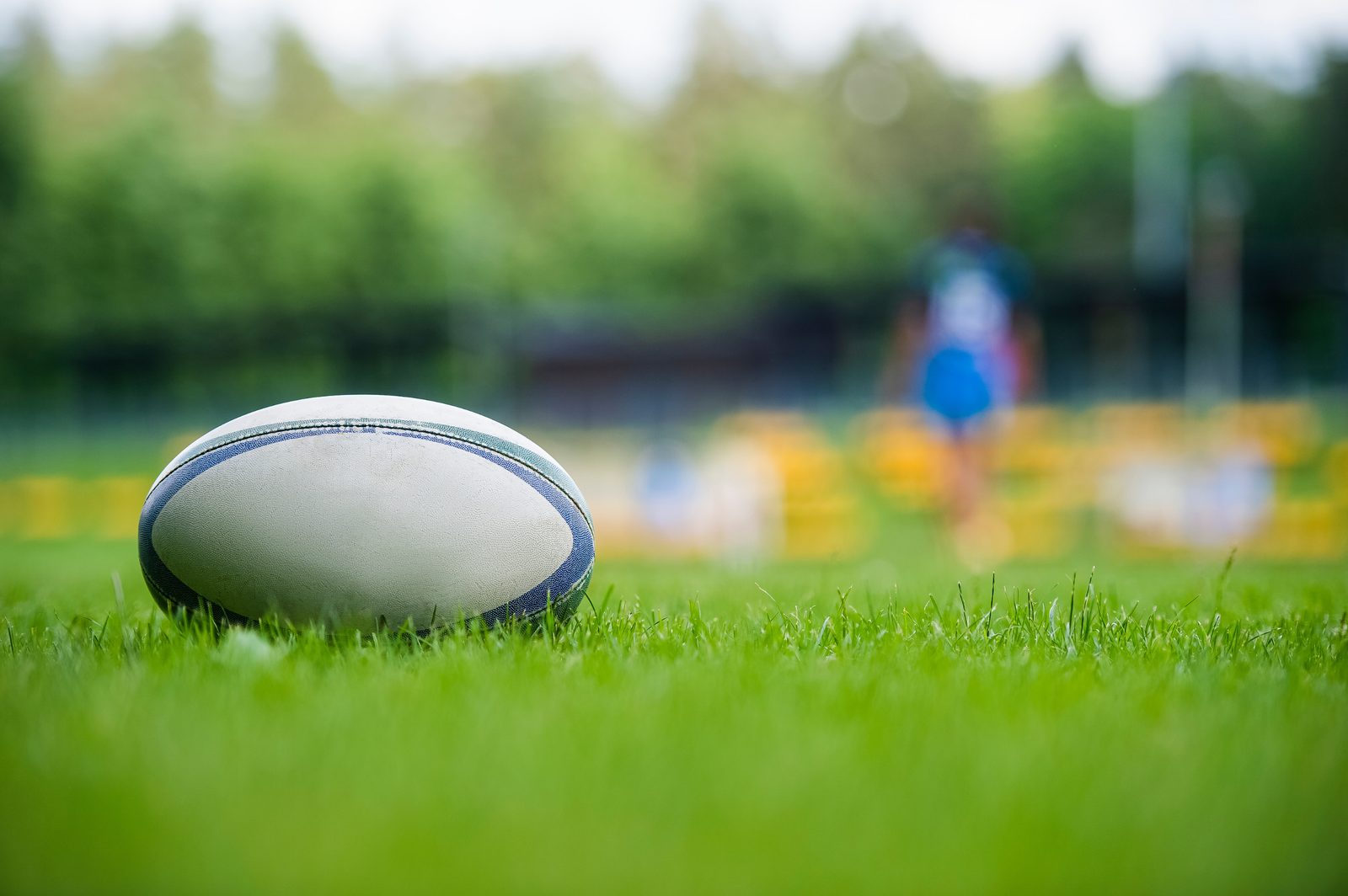 A rugby ball resting on the grass with a training session in the background